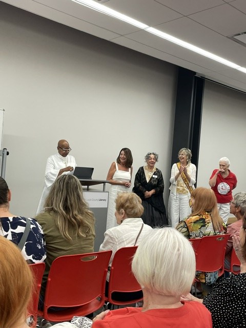 Speakers standing at the front of a St. Louis County Library meeting room, applauded by a seated audience, at the Voices of American HERstory event