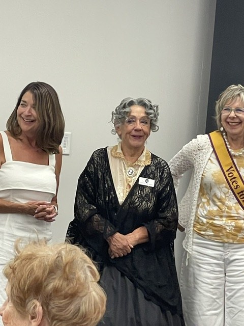 A suffragist reenactor in black lace and a period gown, flanked by a woman in a Votes for Women sash, at the Voices of American HERstory event